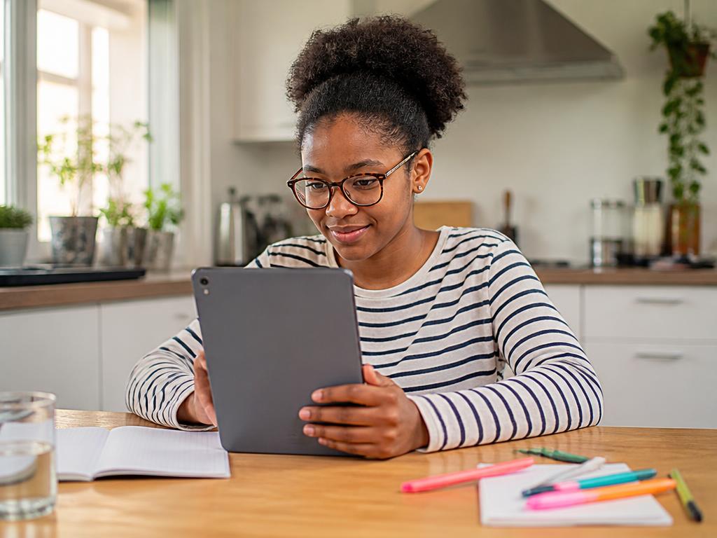 A smiling teenage girl with glasses studying maths on a tablet at a kitchen table